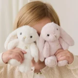 Person Holding Two Fluffy Bunny Plush Toys Up to Their Face One White and One Pale Pink in Front of a Light Background