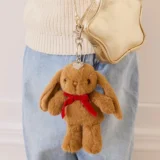 Brown Plush Bunny Keychain with Red Ribbon Hanging from a Metal Clasp Attached to a Gold Handbag Against a Light Background