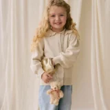 Young Girl with Curly Blonde Hair Wearing a Beige Cardigan and Jeans Smiling While Holding a Small Gold Purse and a Teddy Bear Keychain