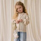Young Girl with Long Curly Hair Wearing a Beige Cardigan and Light Blue Jeans Holding a Small Purse with a Plush Toy Keychain