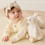 a Baby in Cream Knit Clothing and a Large Headband Bow Lies on the Floor Beside a White Plush Bunny Toy Gazing Upward