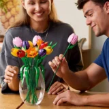 Two People Arrange Bright Plastic Tulips in a Glass Vase on a Wooden Table Smiling As They Work Together