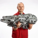 Man in a Red Shirt Holding a Large Gray Lego Spaceship Model in Front of a Plain Background