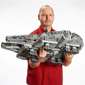 Man in a Red Shirt Holding a Large Gray Lego Spaceship Model in Front of a Plain Background