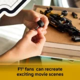 Close up of Hands Assembling a Small Formula 1 Car Model on a Wooden Work Surface with a Yellow Banner Reading 'f1 Fans Can Recreate Exciting Movie Scenes'