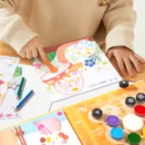 Child Coloring a Vibrant Illustration in a Colorful Art Book with Colored Pencils and Paints Nearby on a Wooden Table