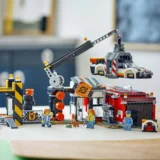 Lego Rescue Scene Crane Lifts a White and Orange Vehicle Above a Red Building with Three Minifigures and a Small Dog Nearby on the Table Inside a Workshop Setting